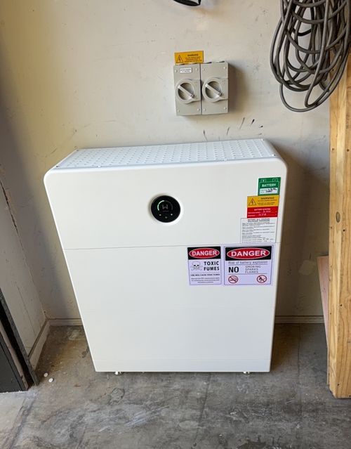 White home battery unit installed on a garage floor against the wall, with isolator switches and warning signs mounted above