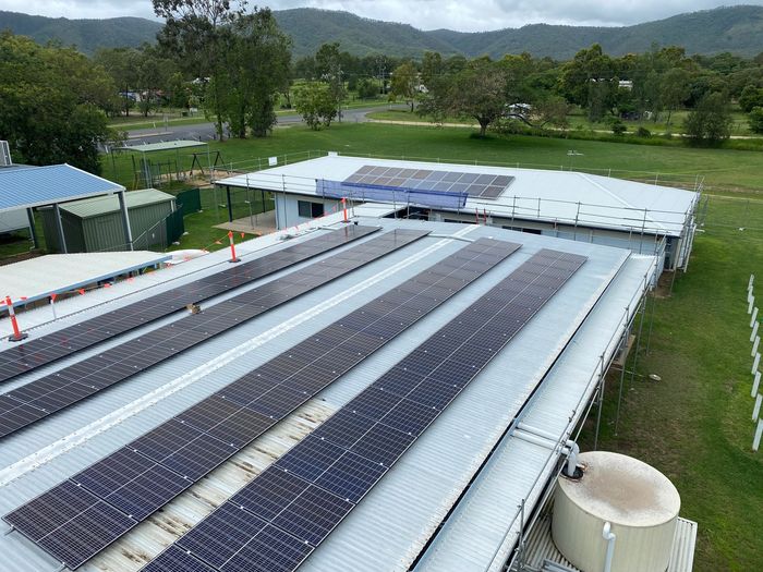 Aerial view of a large solar array on a single-storey building with scaffolding, bushland and hills behind
